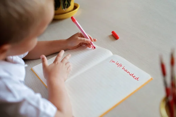 Glad vänsterhänt pojke skriver i pappersboken med sin vänstra hand, internationell vänsterhänt dag — Stockfoto