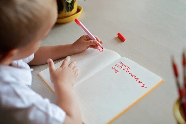 Glad vänsterhänt pojke skriver i pappersboken med sin vänstra hand, internationell vänsterhänt dag — Stockfoto