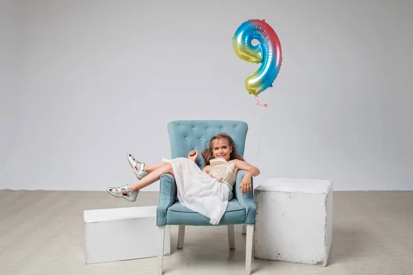 Fille aux cheveux longs en robe de fête s'assoit sur un fauteuil sur fond blanc, anniversaire, plan de studio — Photo