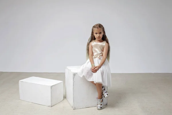穿着节日服装长发的可悲女孩坐在正方形的讲台上，白色的背景，摄影棚的镜头 — 图库照片