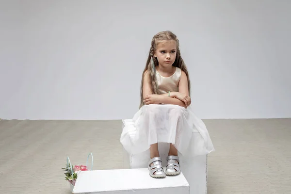 Sad girl with long hair in festive dress sits on square podium, white background, studio shot — Fotografia de Stock