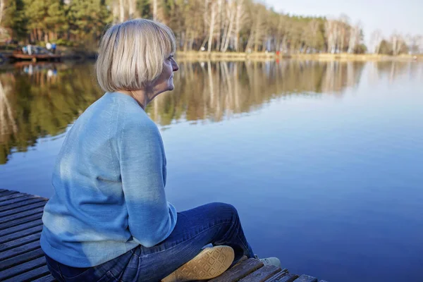 Старшая женщина сидит на берегу озера, отдыхает и наслаждается спокойствием и природой, психическим здоровьем Стоковое Фото