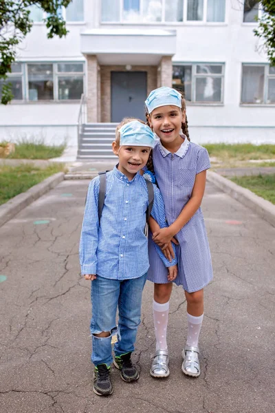 De vuelta a la escuela. Los hermanos con mochilas en máscaras médicas permanecen cerca de las puertas antes del primer día sin conexión —  Fotos de Stock