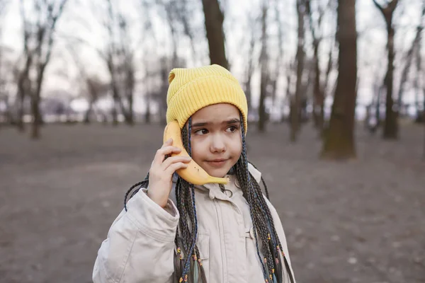 Dziewczyna w żółtym kapeluszu mówi na bananie jak telefon komórkowy w jesiennym lesie, cyfrowy żart detox, styl życia — Zdjęcie stockowe