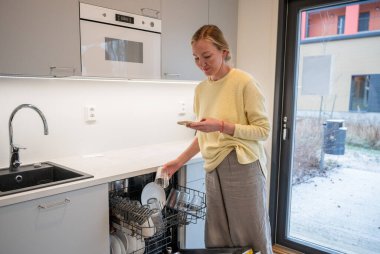 Modern mutfakta akıllı telefon kullanan bir kadın bulaşık makinesini boşaltıyor. Yüksek kalite fotoğraf