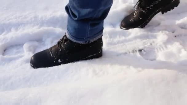 Voeten van een vrouw die in de sneeuw loopt op een besneeuwde dag in zwarte laarzen en blauwe jeans. Zijaanzicht. Wandelen in de besneeuwde stad. — Stockvideo