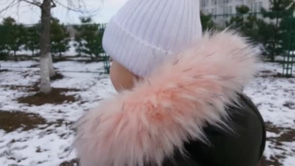 Beyaz şapkalı bir kız karlı şehirde yürüyor. Erimeye başladı, kar erimeye başladı. Kamera hareketliliği arkadan yüze. — Stok video