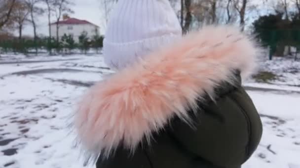 Dívka v bílém klobouku prochází zasněženým městem. Rozmrazilo se, sníh začal tát. Mladá dívka kráčí po zasněžené stezce. Střelba zezadu. — Stock video