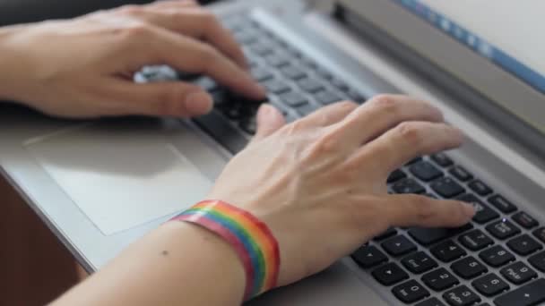 La chica está enviando mensajes de texto en un ordenador portátil, y en su mano es una pulsera con símbolos LGBTQI. — Vídeos de Stock