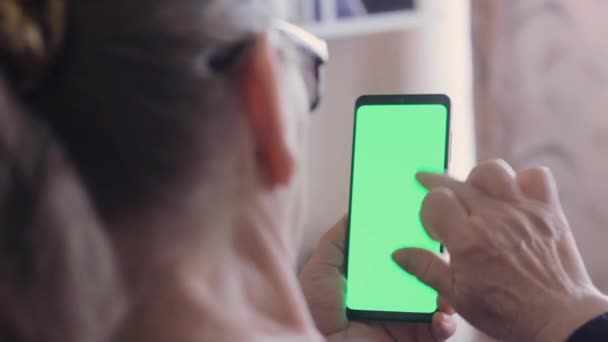 Una anciana con gafas sostiene un teléfono con una pantalla verde en la habitación. Vista posterior de la mujer mayor en el teléfono con pantalla verde para copiar el espacio. — Vídeo de stock