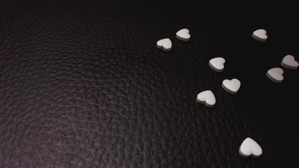 Muitas pílulas em forma de coração branco caem na superfície de couro preto. — Vídeo de Stock