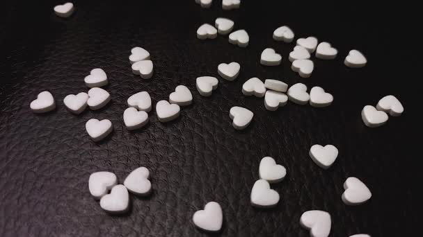 Muchas píldoras blancas en forma de corazón caen sobre la superficie de cuero negro. — Vídeos de Stock
