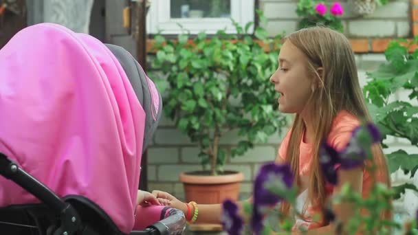 Een mooi tienermeisje met lang stromend haar speelt in de tuin met haar zusje, die in een roze wandelwagen zit. — Stockvideo