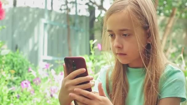 Красивая девочка-подросток с длинными волосами сидит в саду. Ребенок играет по телефону в природе. — стоковое видео