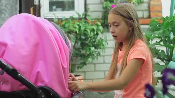 Egy gyönyörű tini lány hosszú hajjal játszik az udvaron a kishúgával, aki egy rózsaszín babakocsiban ül. — Stock videók