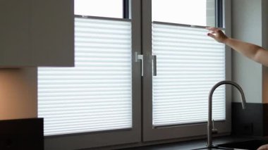 Kafkas bir kadının mutfak penceresinde beyaz jaluziler açması, günışığının modern plisses tonları. Yüksek kaliteli FullHD görüntüler