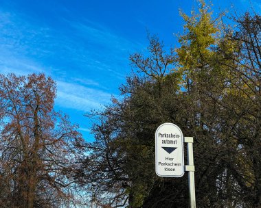 Alman park cezası makinesi Parkscheinotomat açık otoparka, mavi gökyüzüne ve arka plandaki ağaçlara işaret veriyor.