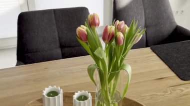 Ahşap bir masadaki cam vazoda parlak renkli laleler, dekoratif mumlar ve modern sandalyeler eşliğinde kapalı bir alanda