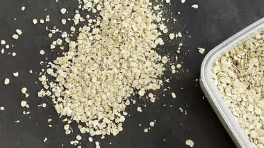 Yulafların açık bir kabın içinden karanlık bir yüzeye döküldüğü görülür. Mutfak ortamında tanelerin dokusunu ve yayılmasını vurgular.