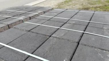 Gri beton kaldırım taşları ahşap paletlerin üzerine istiflenmiş, yeşil kayışlarla güvence altına alınmış, çakıl ve asfalt yolun yanında bir inşaat alanında bulunuyor. Yüksek kaliteli FullHD görüntüler