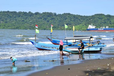Cilacap, Endonezya - 18 Haziran 2025: Cilacap, Central Java 'daki mavi denizden gelen yerel bir tekne, sahil boyunca oynayan turist ve çocuklarla birlikte kumlu plaja yanaşıyor..
