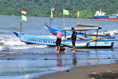 Cilacap, Endonezya - 18 Haziran 2025: Cilacap, Central Java 'daki mavi denizden gelen yerel bir tekne, sahil boyunca oynayan turist ve çocuklarla birlikte kumlu plaja yanaşıyor..