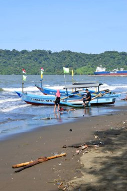 Cilacap, Endonezya - 18 Haziran 2025: Cilacap, Central Java 'daki mavi denizden gelen yerel bir tekne, sahil boyunca oynayan turist ve çocuklarla birlikte kumlu plaja yanaşıyor..