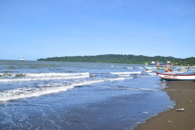 Cilacap, Endonezya - 18 Haziran 2025: Cilacap, Central Java, Endonezya 'da geniş bir kumsal manzarası, geleneksel balıkçı tekneleri, kıyıda bir hindistan cevizi ve uzak bir deniz feneri.