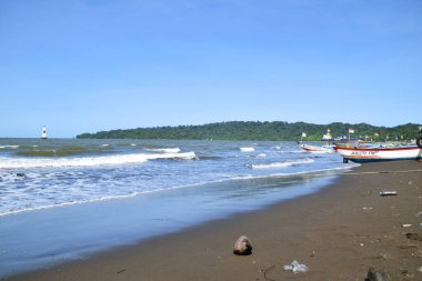 Cilacap, Endonezya - 18 Haziran 2025: Cilacap, Central Java, Endonezya 'da geniş bir kumsal manzarası, geleneksel balıkçı tekneleri, kıyıda bir hindistan cevizi ve uzak bir deniz feneri.