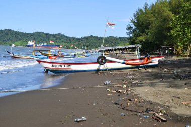 Cilacap, Endonezya - 18 Haziran 2025: Cilacap, Central Java, Endonezya 'da geniş bir kumsal manzarası, geleneksel balıkçı tekneleri, kıyıda bir hindistan cevizi ve uzak bir deniz feneri.