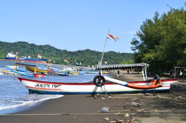 Cilacap, Endonezya - 18 Haziran 2025: Cilacap, Central Java, Endonezya 'da geniş bir kumsal manzarası, geleneksel balıkçı tekneleri, kıyıda bir hindistan cevizi ve uzak bir deniz feneri.