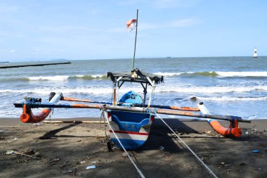 Cilacap, Endonezya - 18 Haziran 2025: Cilacap, Central Java, Endonezya 'da bir plajın koyu kumlu kıyılarına demirlemiş geleneksel bir balıkçı teknesinin okyanus dalgalarıyla arka görüntüsü.