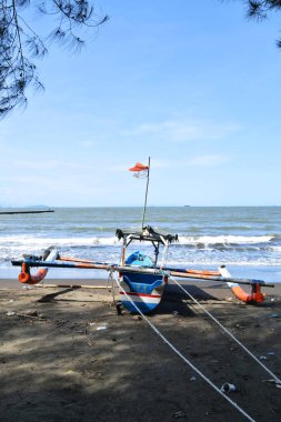 Cilacap, Endonezya - 18 Haziran 2025: Cilacap, Central Java, Endonezya 'da bir plajın koyu kumlu kıyılarına demirlemiş geleneksel bir balıkçı teknesinin okyanus dalgalarıyla arka görüntüsü.