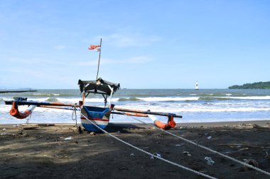 Cilacap, Endonezya - 18 Haziran 2025: Cilacap, Central Java, Endonezya 'da bir plajın koyu kumlu kıyılarına demirlemiş geleneksel bir balıkçı teknesinin okyanus dalgalarıyla arka görüntüsü.