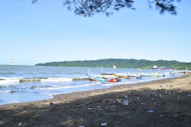 Cilacap, Endonezya - 18 Haziran 2025: Plastik çöp ve molozlarla dolu geniş bir kumsal manzarası, Cilacap, Central Java, Endonezya kıyıları boyunca sıralanmış geleneksel balıkçı tekneleri.