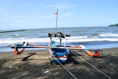 Cilacap, Endonezya - 18 Haziran 2025: Cilacap, Central Java, Endonezya 'da bir plajın koyu kumlu kıyılarına demirlemiş geleneksel bir balıkçı teknesinin okyanus dalgalarıyla arka görüntüsü.
