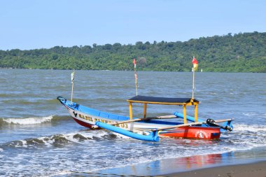 Cilacap, Endonezya - 18 Haziran 2025: Okyanus dalgaları ve Cilacap, Central Java 'da yeşil tepe manzaralı kumlu bir plaja demirlemiş renkli geleneksel bir balıkçı teknesi.