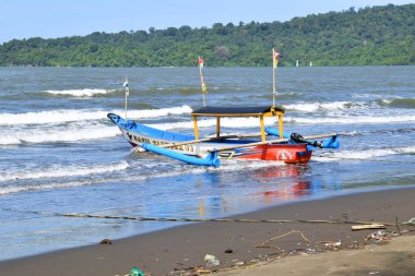 Cilacap, Endonezya - 18 Haziran 2025: Okyanus dalgaları ve Cilacap, Central Java 'da yeşil tepe manzaralı kumlu bir plaja demirlemiş renkli geleneksel bir balıkçı teknesi.