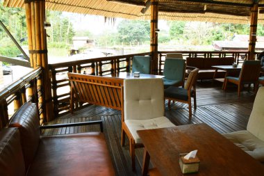 Tahta masalar, minderli sandalyeler ve balkondan doğal manzaralı geleneksel bir bambu yapı restoranının iç manzarası.