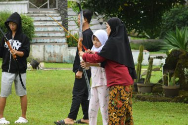 Kuningan, Endonezya - Temmuz 06, 2025: Bir tesettürlü genç kız, bir eğitmen ve diğer çocuklarla Endonezya 'nın Kuningan kentindeki açık yeşil bir alanda okçuluk yapıyor..