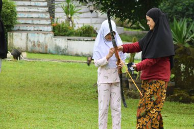 Kuningan, Endonezya - Temmuz 06, 2025: Batik etekli bir anne, Endonezya 'nın Kuningan kentindeki yeşil bir tarlada okçuluk yaparken tesettürlü genç kızına yay tutmasında yardım ediyor.