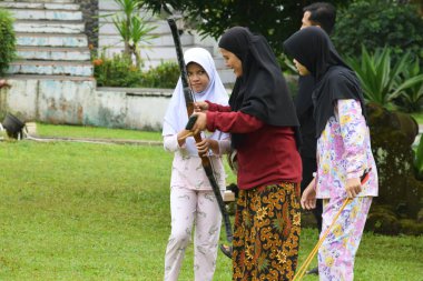 Kuningan, Endonezya - Temmuz 06, 2025: Batik etekli bir anne, Endonezya 'nın Kuningan kentindeki yeşil bir tarlada okçuluk yaparken tesettürlü genç kızına yay tutmasında yardım ediyor.