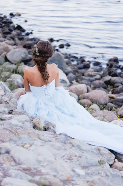 Morena en un exuberante vestido azul con hermoso pelo sentado en el río Banco grandes stones.her se encuentra en el jardín —  Fotos de Stock