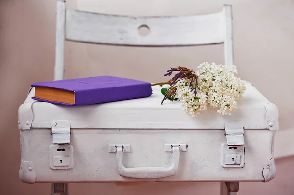 White painted suitcase on a light background, with the white lilac and purple book.