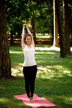 Young beautiful red-haired girl does yoga in Park on green background. .
