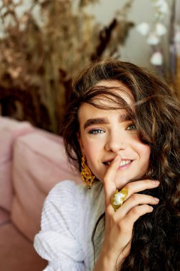 Güzel gözleri, kıvırcık saçları ve kalın kaşları olan bir model. Gülümseyin, parmaklarınızı dudaklarınıza yaklaştırın. Moda ve tarz
