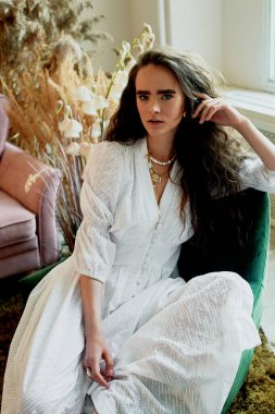 Güzel model beyaz pamuklu bir elbiseyle kadife bir sandalyede oturur. Tarz, moda ve güzellik