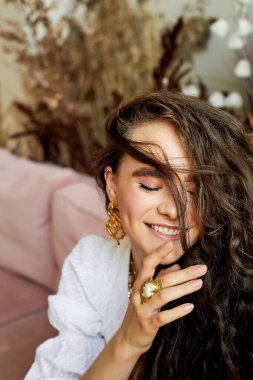 Güzel gözleri, kıvırcık saçları ve kalın kaşları olan bir model. Gülümseyin, parmaklarınızı dudaklarınıza yaklaştırın. Moda ve tarz