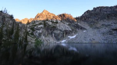 Idahos Sawtooth Dağları 'ndaki Trail Creek Gölü üzerinde gün batımı zamanı. Altın ışık engebeli tepeleri ısıtır. Akşamüstü gökyüzünün altındaki sakin alp gölünde yansımalar parıldar..