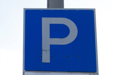 Üzerinde beyaz 'P' işareti olan kare mavi bir park işareti gri bir direğe, parlak beyaz bir gökyüzüne monte edilmiştir..